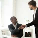Standing business man shaking another business man's hand who's sitting down in an office setting