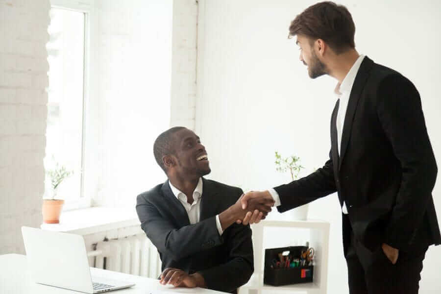 Standing business man shaking another business man's hand who's sitting down in an office setting