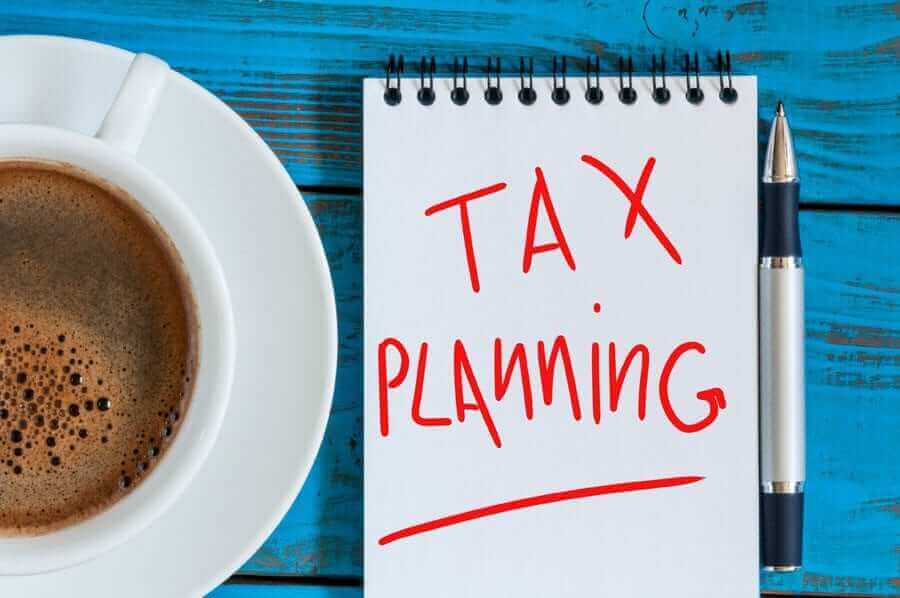Notepad with "Tax Planning" written in red and underlined next to coffee cup on blue desk