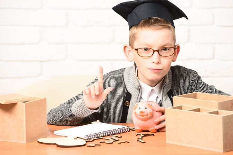 Boy with glasses and graduation hat holding piggy bank and one finger up