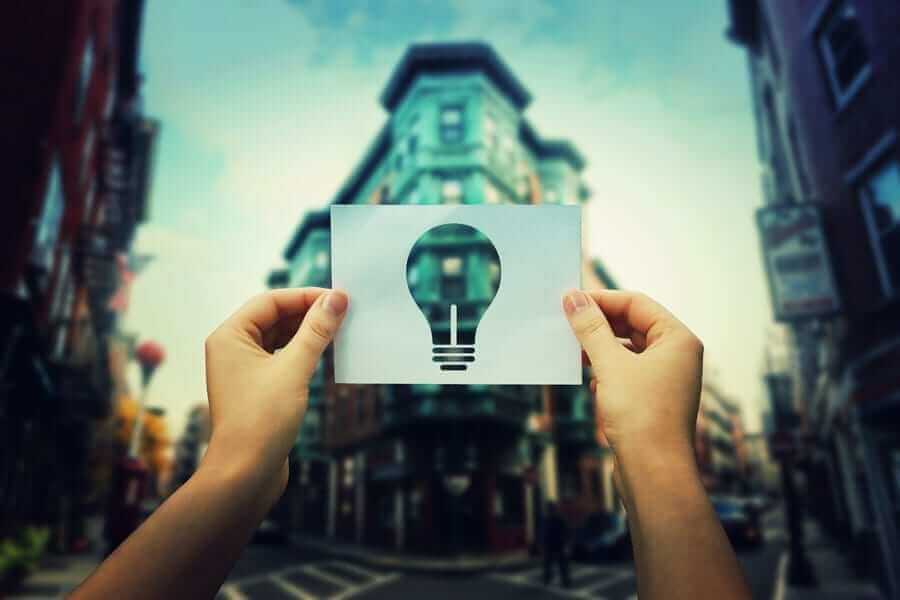 Hands in foreground holding up paper with light bulb shape cut out in front of building
